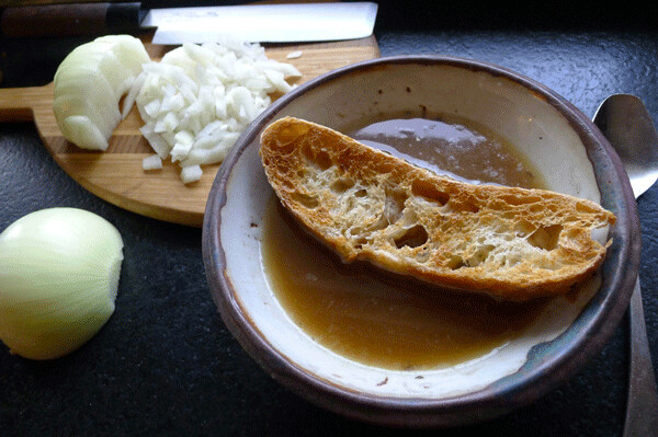 A crusted bowl of French onion soup, with minced onions in the background. Ari LeVaux